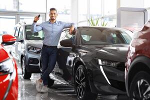 Stylish arab man holding key from automobile and smiling, buying brand new sports car in showroom. Happy middle-eastern guy standing by nice black auto and showing automatic key at camera photo