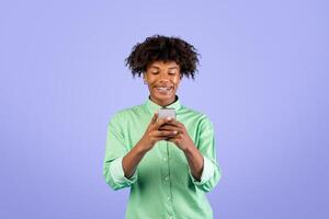People and gadgets. Happy black guy using smartphone, smiling, browsing internet and scrolling social media news feed standing over blue studio background photo
