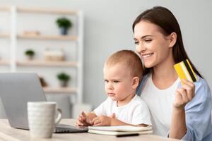 Happy Young Woman Holding Toddler Baby And Making Online Shopping At Home, Smiling Millennial Mother With Infant Child Using Laptop And Credit Card, Enjoying Purchasing In Internet, Copy Space photo