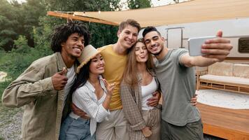 Portrait of happy diverse friends posing near their camper van in countryside, making different gestures, taking selfie on smartphone, panorama. Cheerful young people having camping trip in summer photo