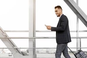 Handsome arab guy using smartphone while walking with suitcase at airport terminal, young middle eastern man browsing mobile internet on cellphone while going to flight boarding, side view photo