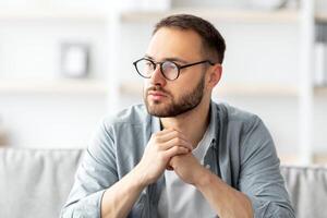 Serious young Caucasian guy sitting on couch in front of laptop computer at home. Thoughtful millennial man daydreaming while working online, using portable pc indoors photo