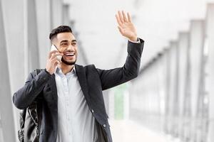 Hello. Happy Arab Man Waving Hand And Talking On Cellphone While Walking In Airport, Handsome Smiling Middle Eastern Guy Going Out Of Terminal After Flight Arrival And Greeting Somebody, Free Space photo