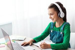 Studying At Home. Cheerful schoolgirl doing homework and typing on keyboard, sitting at table using laptop, looking at screen, writing essay or making test, taking notes in textbook, free copy space photo