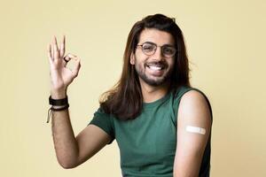 Cheerful long-haired indian guy showing okay gesture and smiling at camera, got vaccinated against COVID-19, having adhesive medical band on shoulder, beige studio background, copy space photo