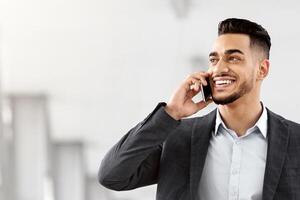 Young Middle Eastern Businessman Walking With Suitcase In Airport And Talking On Cellphone, Handsome Arab Man In Smart Casual Clothes Enjoying Phone Call While Going For Flight Gate In Terminal photo
