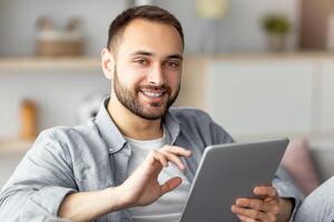 Online life concept. Happy young man using tablet computer, working or studying from home. Handsome Caucasian guy taking part in webinar or internet conference, having business meeting photo