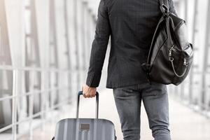 Rear View Of Unrecognizable Male Traveller With Backpack And Suitcase Walking In Airport, Back Shot Of Young Man Passing Terminal Corridor On His Way To Flight Boarding Gate, Copy Space photo