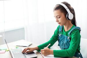 Educational Lecture Concept. Portrait of diligent teen girl in wireless headphones sitting at table using laptop, writing in copybook taking notes in notebook, listening to teacher, free copy space photo