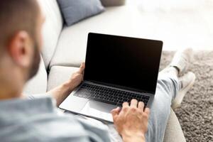 Unrecognizable young man using laptop on comfy sofa at home, mockup for website design on screen. Millennial guy holding portable pc with copy space, working or studying online photo