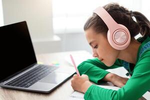 Side view profile of focused little girl writing down information watching webinar on laptop with empty screen or taking notes in her diary, wearing wireless headphones, pc with free space mock up photo