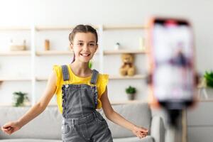 Portrait of young teenage blogger girl dancing at camera shooting tutorial using phone on tripod at home, creating trendy content on mobile application to share on social media, selective focus photo