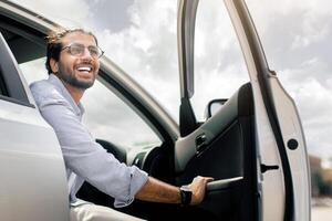Positive young middle-eastern man going out the car, low angle photo, copy space. Happy arab guy open automobile door and smiling, getting in. Auto test-drive, leasing, buying, renting concept photo