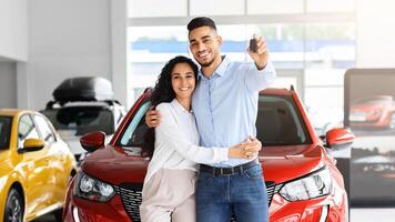 Happy beautiful loving muslim family young arab man and long-haired woman buying new car in salon, standing over red luxury automobile, hugging and showing key from their new auto, copy space photo