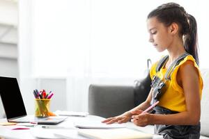 Online Education Concept. Profile side view of school girl studying at home, writing in her copybook or diary, doing homework or attending webinar using laptop with empty blank screen on table photo