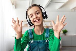 E-learning. Portrait of smiling school girl making call with teacher and classmates, using pc. Cheerful student wearing wireless headphones talking and waving hands, web camera point of view photo