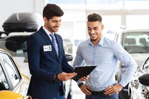 Attractive middle-eastern man manager showing chart with car specifications and smiling while having conversation with handsome wealthy man, helping with choosing new automobile, copy space photo
