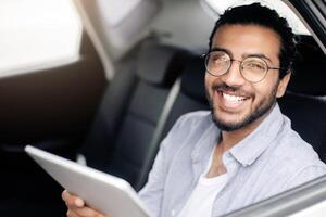 Cheerful indian guy using digital tablet while taking cab, copy space. Smiling middle-eastern young man having car trip, sitting on back seat and reading blog on pad. Transportation service concept photo