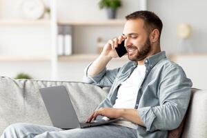 Attractive young guy talking on smartphone and using laptop computer on couch at home office. Handsome male businessman having phone conversation with client, working on portable pc photo