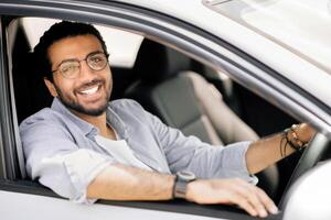 Car leasing, buying, retail, renting concept. Portrait of happy arab guy in casual outfit sitting in brand new auto, placing hand on wheel, cheerfully smiling at camera, side view, copy space photo
