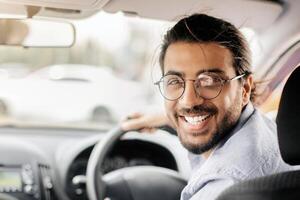 Cheerful middle-eastern young man taxi driver looking at back seat and smiling at camera, closeup photo, copy space. Happy arab guy enjoying his brand new automobile, making test-drive photo