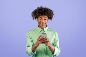 Cool app. Happy african american guy using smartphone, browsing new application or messaging with friends online, standing over blue studio background, copy space photo