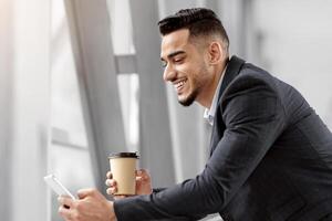 Happy Middle Eastern Businessman Using Smartphone And Having Coffee While Waiting In Airport Terminal, Handsome Arab Man Browsing Internet On Cellphone Or Answering Emails And Enjoying Takeaway Drink photo