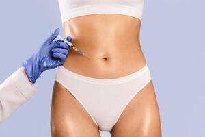 Beauty Procedure For Body. Slim woman having lipolysis treatment at beauty salon, cropped closeup. Plastic surgeon in gloves making injection at belly area for unrecognizable lady, blue background photo