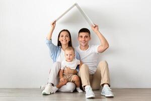 Real Estate Concept. Happy Family With Little Toddler Child Holding Symbolic Cardboard Roof Above Head, Smiling Young Spouses And Their Infant Son Dreaming About Moving To New Home, Copy Space photo