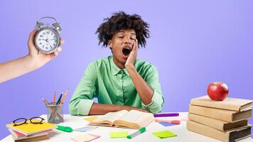 Tired african american male student yawning, sitting at desk with lot of books and preparing for exams against blue studio background. Hand with alarm clock. Deadline and overworking concept. photo