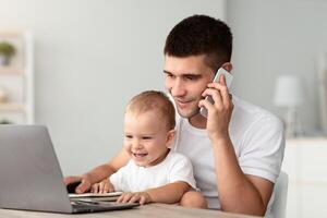 Handsome Young Father Talking On Cellphone While Baby Using Laptop At Home, Freelancer Guy Sitting At Desk With Computer, Working Remotely By Cellphone And Taking Care About Toddler Son, Copy Space photo