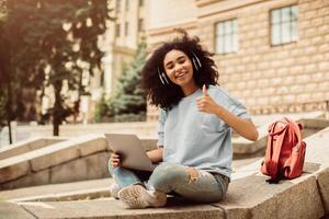 Black Teenager Girl Learning Online Using Laptop And Showing Like Gesture Smiling To Camera Approving E-Learning And Distance Study Sitting Outside. Student With Computer Gesturing Thumbs Up photo
