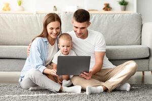 Happy Young Parents And Their Toddler Baby Using Laptop Together At Home While Relaxing On Floor In Living Room, Millennial Family Of Three Shopping Online Or Browsing Internet On Computer photo