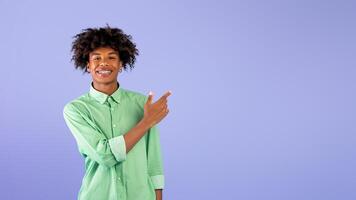 Look there. Happy african american teen guy pointing aside with index finger at empty space, standing over blue background, studio shot, panorama photo