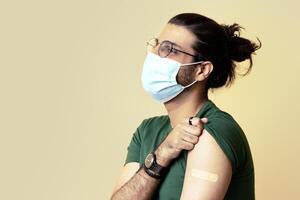 Long-haired indian man in protective face mask got vaccinated against COVID-19, showing shoulder with plaster on, sitting on chair over beige studio background, looking at copy space, panorama photo