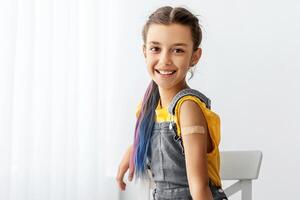 Vaccination, Protection. Happy Adolescent Girl Showing Vaccinated Arm With Adhesive Bandage Plaster On Shoulder After Getting Corona Virus Covid Vaccine In Clinic Sitting On Chair. Antiviral Injection photo