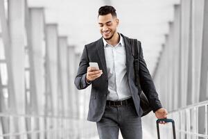 Online Check In. Handsome Arab Man Walking With Suitcase In Airport And Using Smartphone, Smiling Young Middle Eastern Guy Browsing Internet On Cellphone While Going To Flight Gate, Copy Space photo