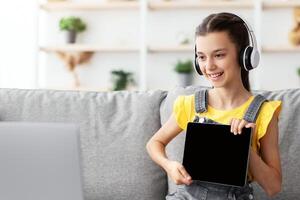 Distance Education. Smiling girl having call with teacher, holding digital tablet with blank empty screen showing done task result using laptop. Child studying remotely photo