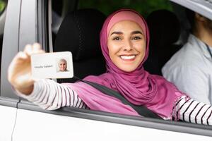 Joyful Islamic Lady In Hijab Demonstrating Driver Licence At Camera While Sitting In Car Next To Instructor, Cheerful Muslim Female In Headscarf Celebrating Passing Exam In Drive School, Closeup photo