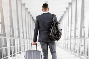 Young Man In Casual Clothes Walking With Luggage In Airport Terminal, Rear View Of Unrecognizable Male Traveller Carrying Suitcase And Backpack While Going To Flight Boarding Gate, Copy Space photo