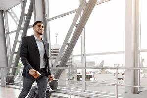 Ready For Journey. Smiling Handsome Middle Eastern Man Walking With Suitcase In Airport, Young Arab Guy Carrying Luggage Passing Terminal Corridor, Going To Boarding Gate, Panorama With Copy Space photo