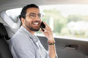 Positive young indian man freelancer working online while sitting at backseat in taxi, typing on laptop with blank screen and having phone conversation, mockup, looking at copy space, side view photo