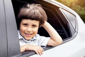 Closeup Shot Of Cheerful Little Boy Looking Out Of Car Window While Having A Ride, Portrait Of Adorable Small Male Child Enjoying Automobile Travels, Sitting Inside Of Auto And Smiling At Camera photo