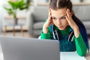 Information Overflow And Overload. Little female student sitting at desk touching temples grabbing head looking at pc computer screen, bored and unmotivated girl feeling tired of unfinished homework photo