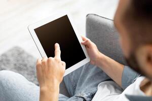 Advertisement concept. Millennial man using tablet computer with blank black screen, browsing website or demonstrating new app, sitting on couch at home. Mockup for your design photo