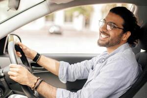 Handsome long-haired indian guy with glasses driving brand new car, looking at rear view mirror and smiling, side view, copy space. Cheerful middle-eastern young man enjoying his new automobile photo