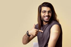 Handsome millennial long-haired indian man got vaccinated against COVID-19, pointing at shoulder with plaster on and smiling at camera, sitting on chair over beige studio background, copy space photo