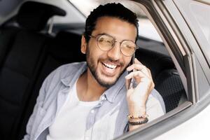 Positive handsome arab guy taking cab, having phone conversation while going to job or home, middle-eastern young man sitting at taxi back seat and talking on smartphone, copy space photo