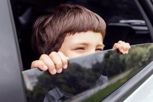 Cheerful little boy looks out the open window and enjoys ride with parents in car in summer, outdoor, free space. Journey with family at weekend, vacation together, road and travel by auto, close up photo