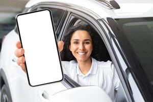 Positive millennial middle eastern woman sitting in brand new white car, showing smartphone with empty screen and smiling at camera, mockup, renting auto on Internet. Car dealership online photo